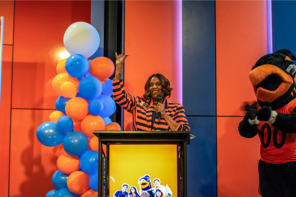 LT stands at podium holding up a Birds Up handsign with a blue and orange balloon tower on the left and Rowdy stands at the right